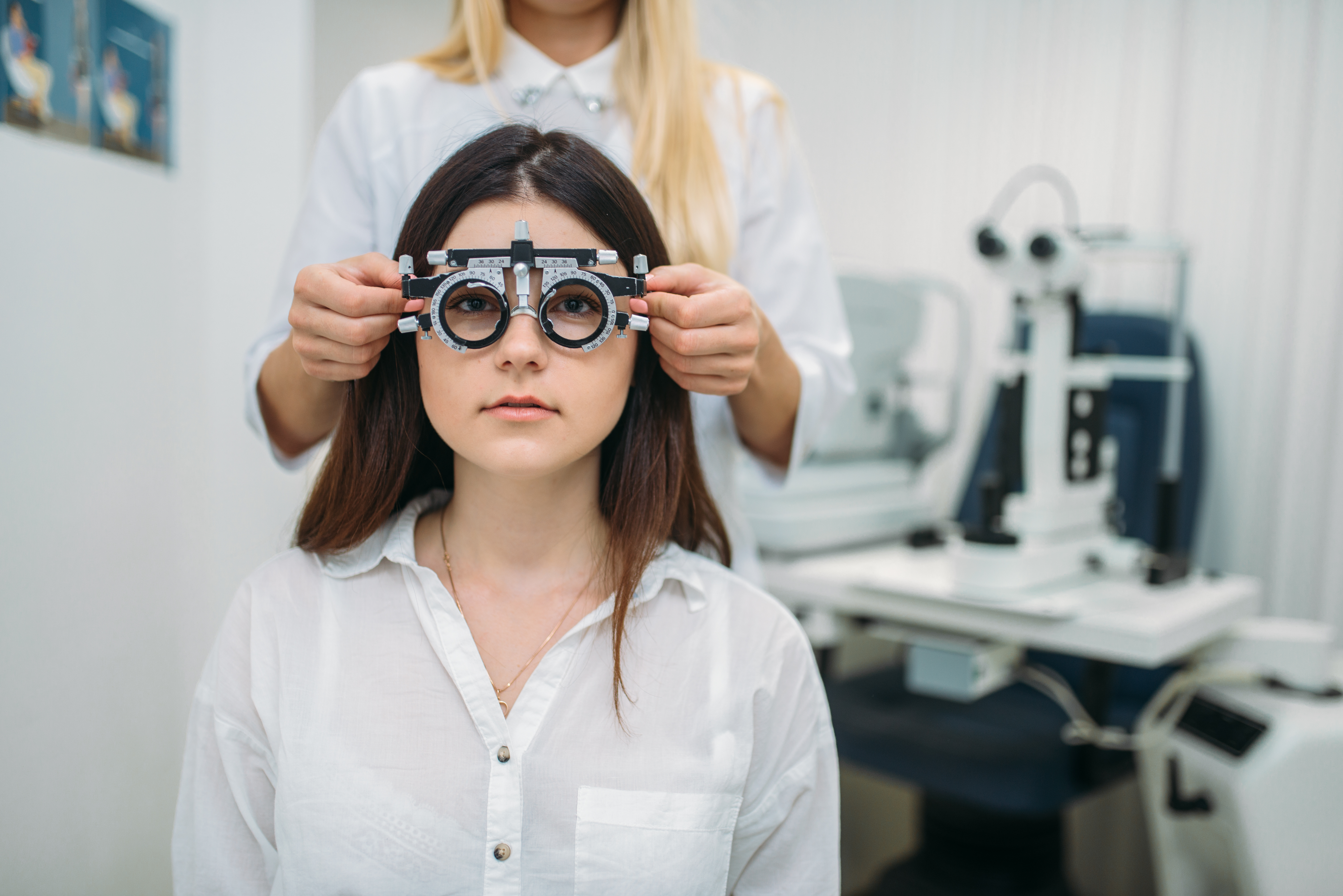 Frau sitzt vor optischem Gerät, während eine andere Person ihr eine Sehhilfe anpasst