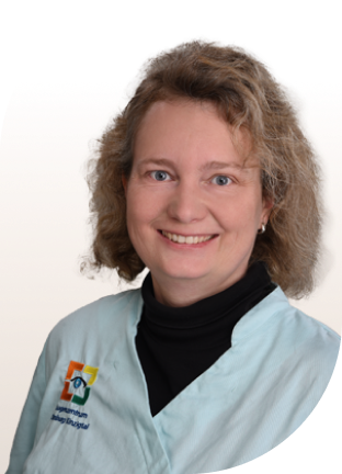 Person mit schulterlangem, lockigem Haar trägt hellblaue Arbeitskleidung mit schwarzem Rollkragenpullover und Ohrring.